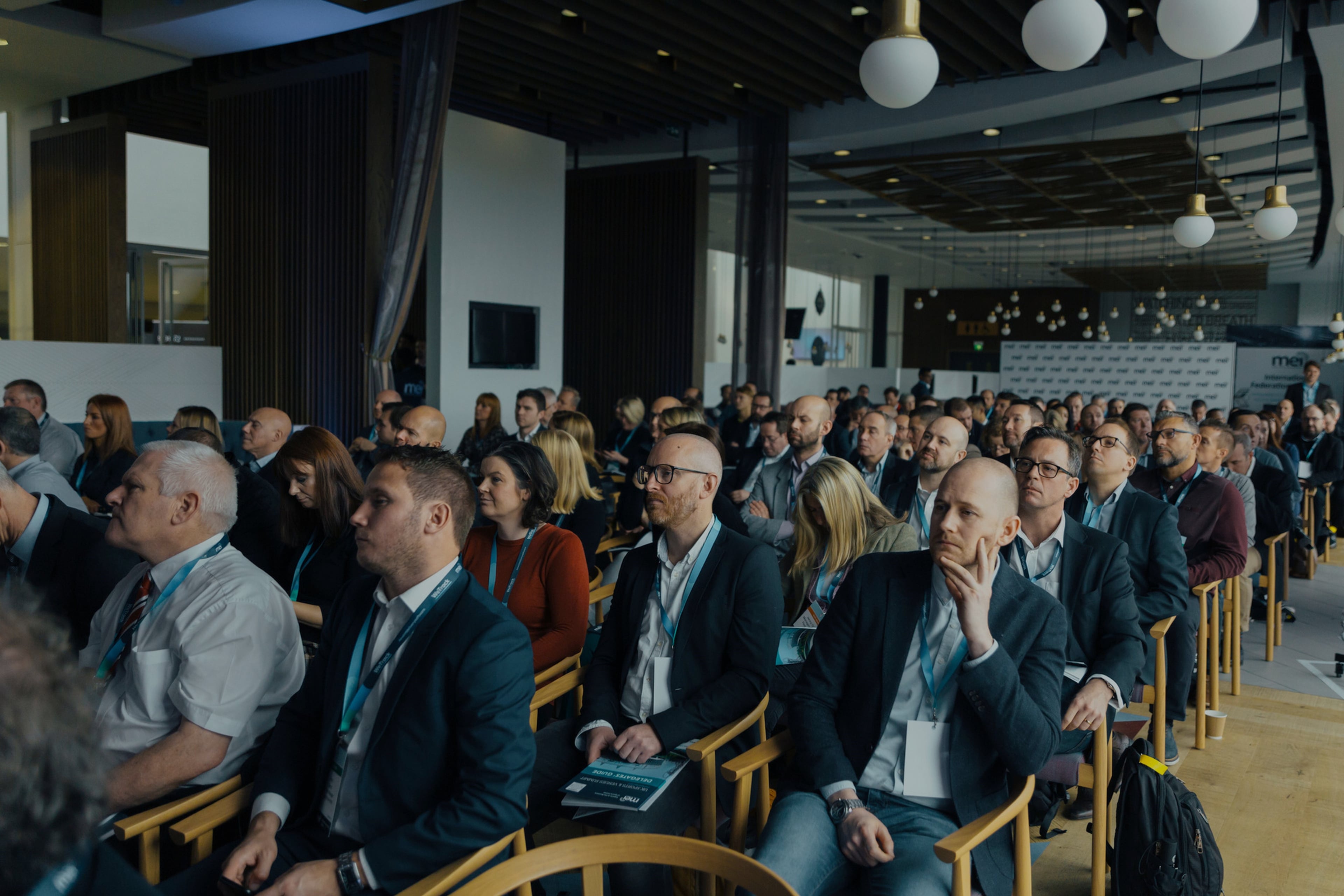 Deltagere på konference sidder på træstole og lytter til præsentation i en moderne sal